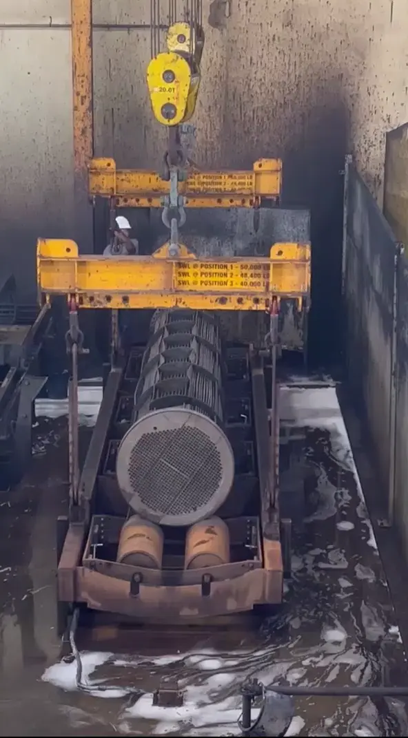 A worker oversees a crane lifting a large heat exchanger from a wet, industrial cleaning area.